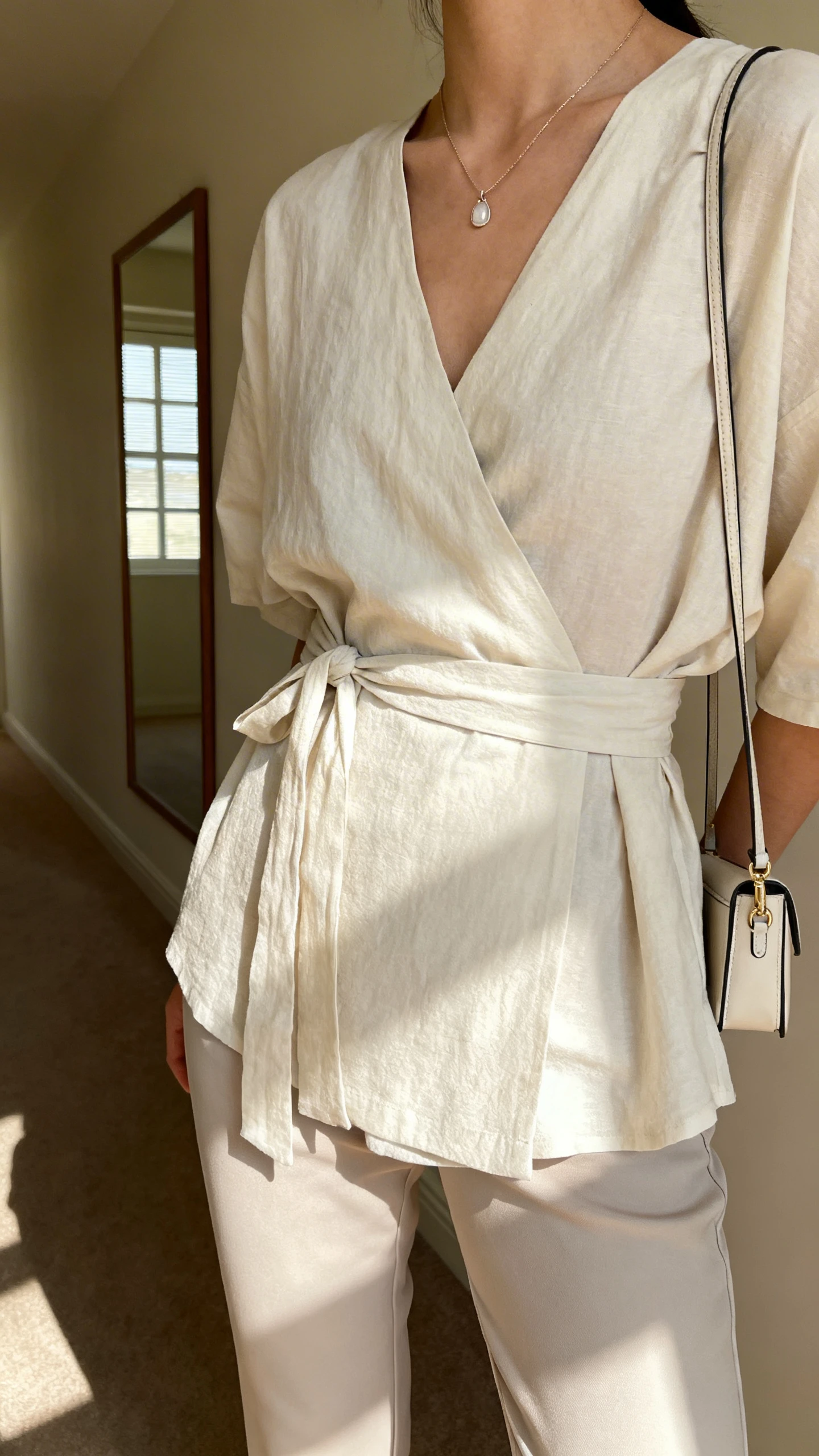 iPhone back/side view of a woman wearing an easy wrap top with tie at the waist over slim trousers, delicate pendant necklace and small crossbody, face not visible, hallway mirror, afternoon window light, iPhone photo quality.
