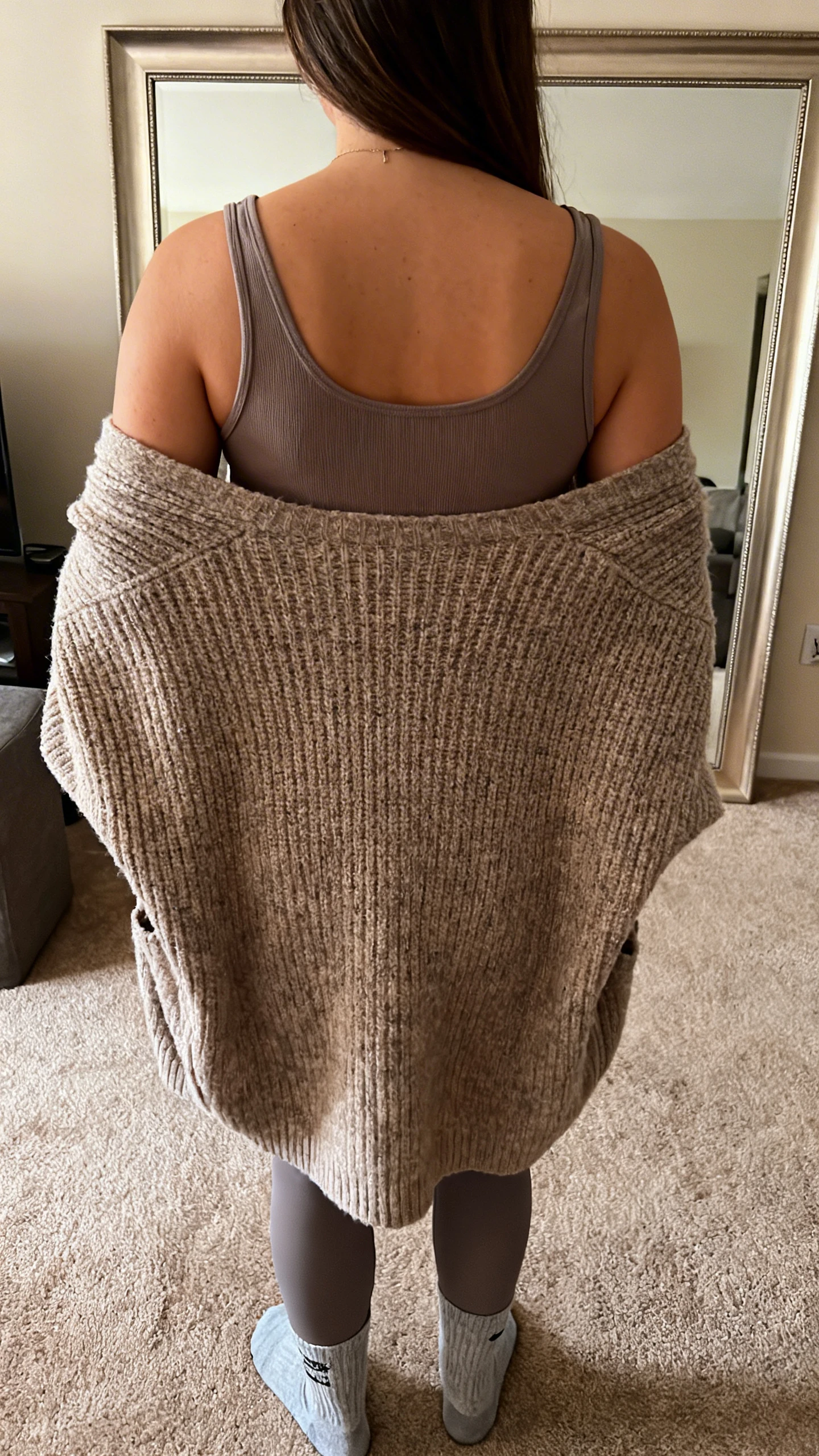 iPhone back-view photo of a woman in a cozy cocoon cardigan layered over a fitted tank with leggings and socks, face not visible, living room mirror, warm indoor lighting, iPhone photo quality.