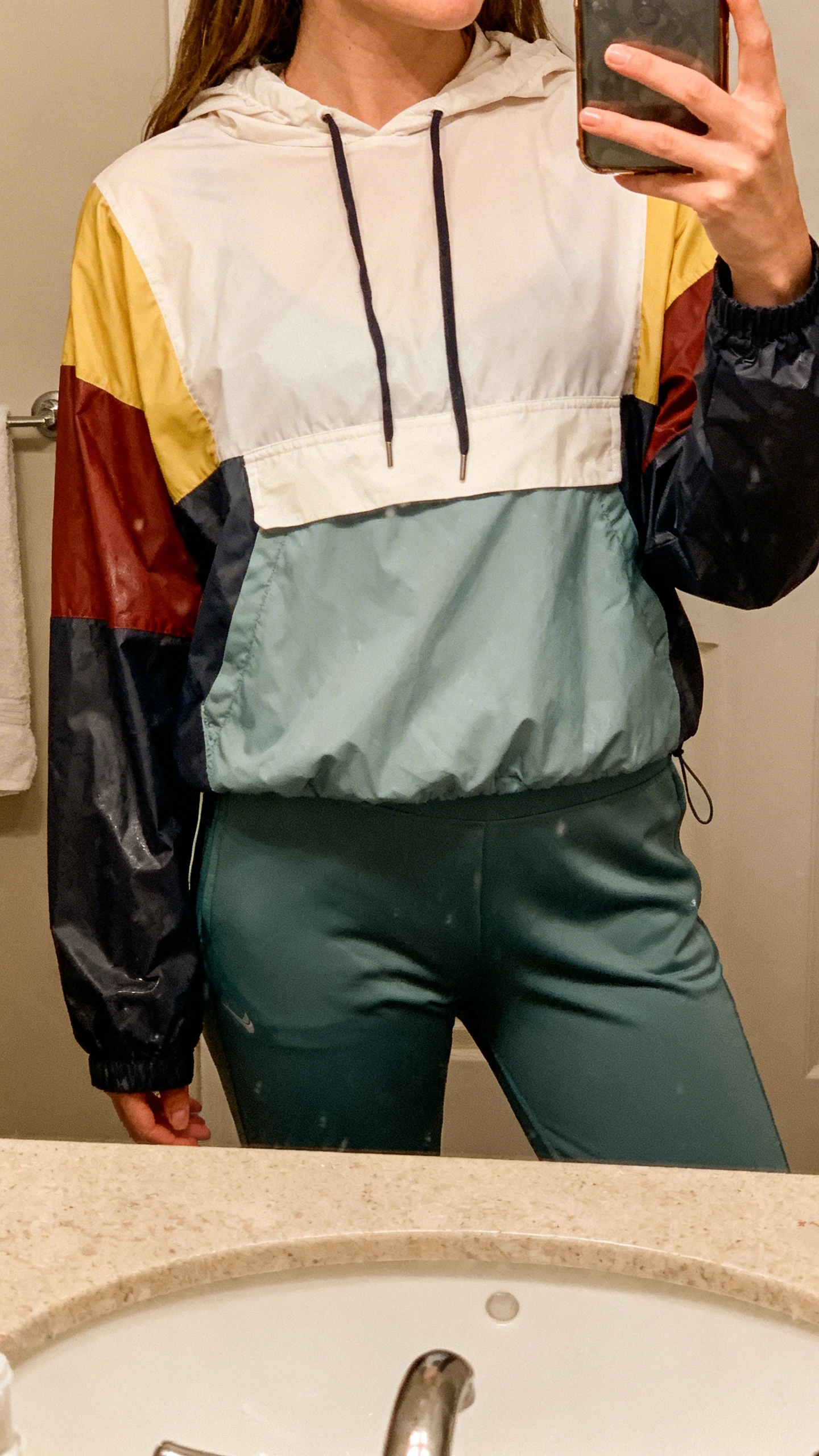 iPhone bathroom mirror selfie of a woman wearing a rain-ready raglan hoodie with color-block panels and slim joggers, face not visible, cropped at shoulders, indoor lighting, iPhone photo quality.
