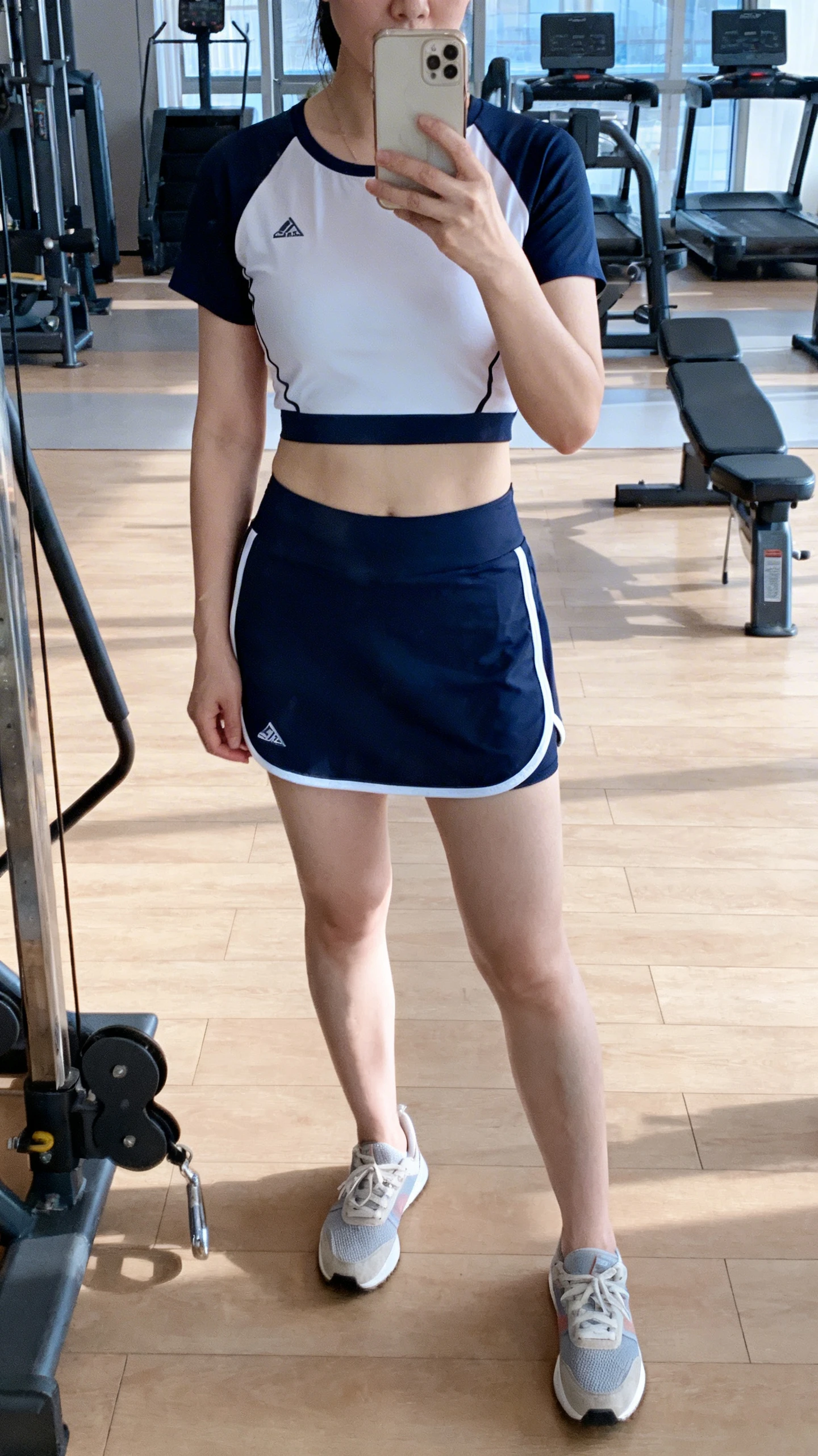 iPhone mirror selfie of a woman in a sporty skort set with contrast binding and a matching athletic top, low-profile trainers, face not visible, gym mirror, bright indoor lighting, iPhone photo quality.
