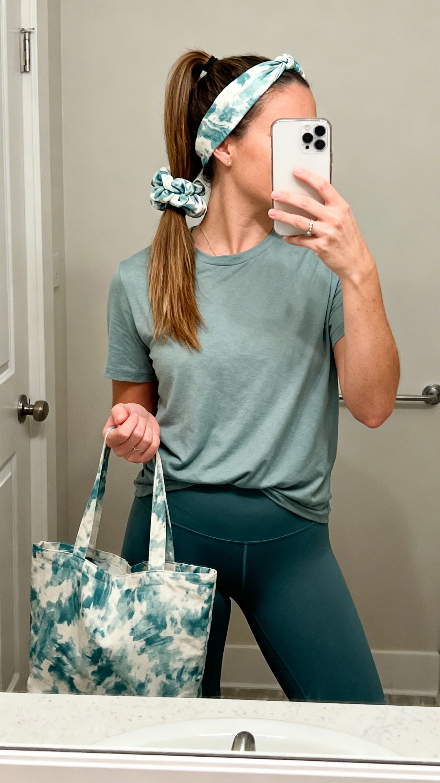 iPhone mirror selfie of a woman wearing a matching scrunchie in her ponytail and a fabric headband, styled with a basic tee and leggings while holding a coordinating tote, face not visible, cropped at neck, bathroom mirror, natural indoor lighting, iPhone photo quality.