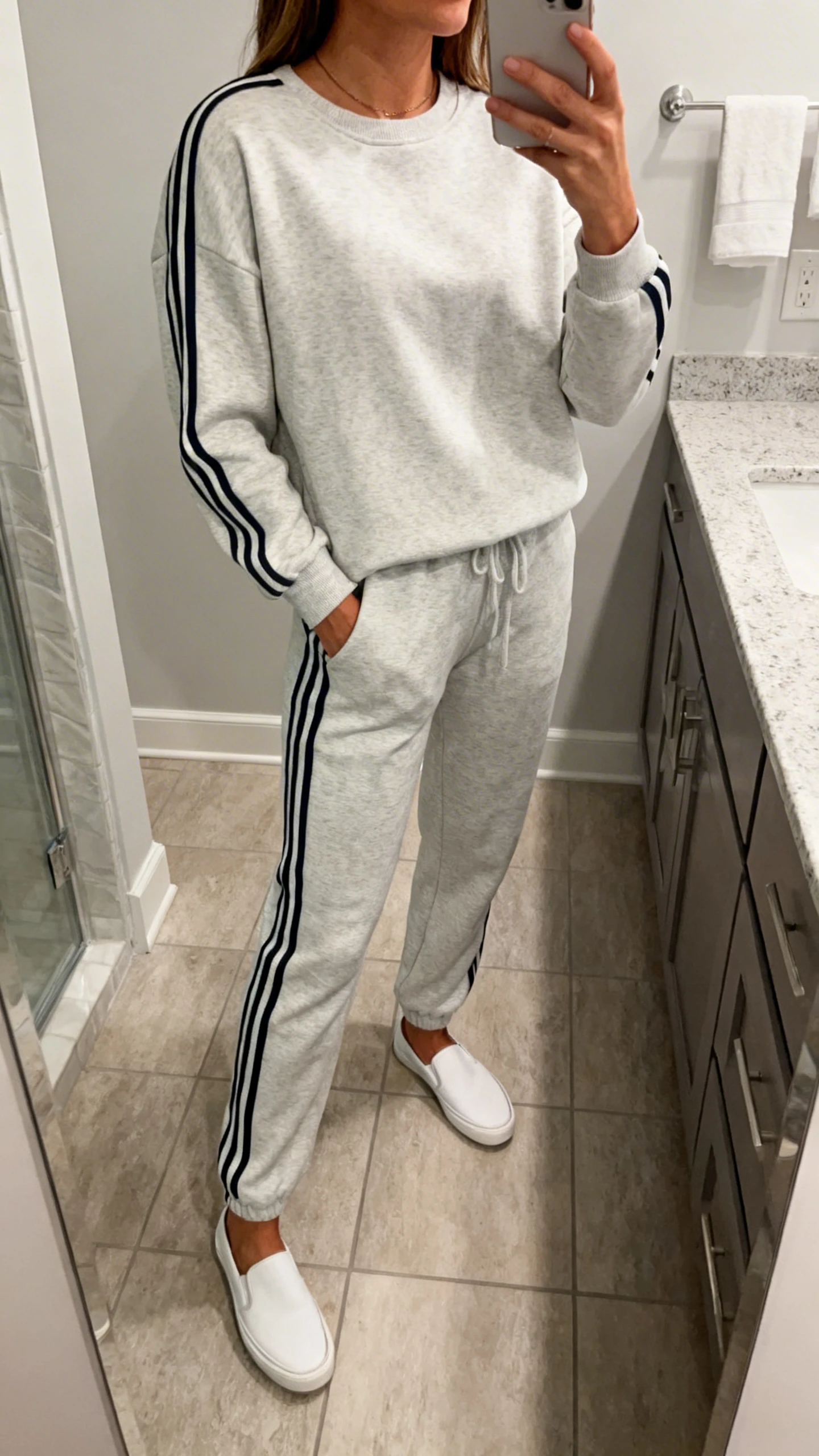 iPhone mirror selfie of a woman wearing a weekend jogger set with side stripes and a matching pullover, slip-on sneakers, face not visible, cropped at neck, bathroom mirror, indoor lighting, iPhone photo quality.