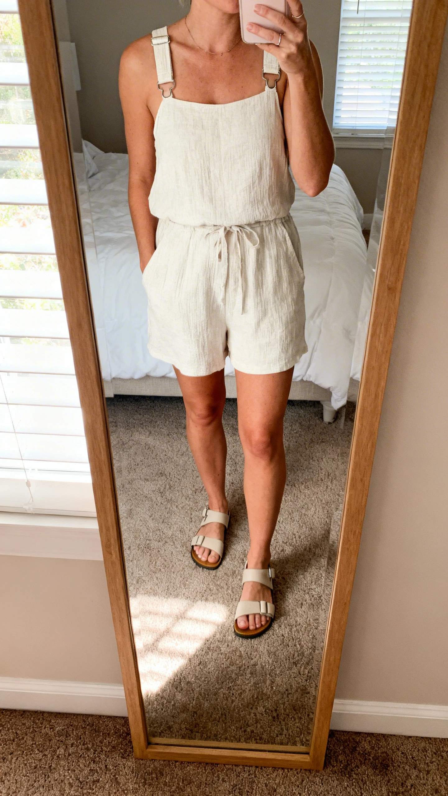 iPhone mirror selfie of a woman wearing an easy-peasy playdate romper with grow-with-me adjustable straps and simple sandals, face not visible, cropped at neck, bedroom mirror, soft morning window light, iPhone photo quality.