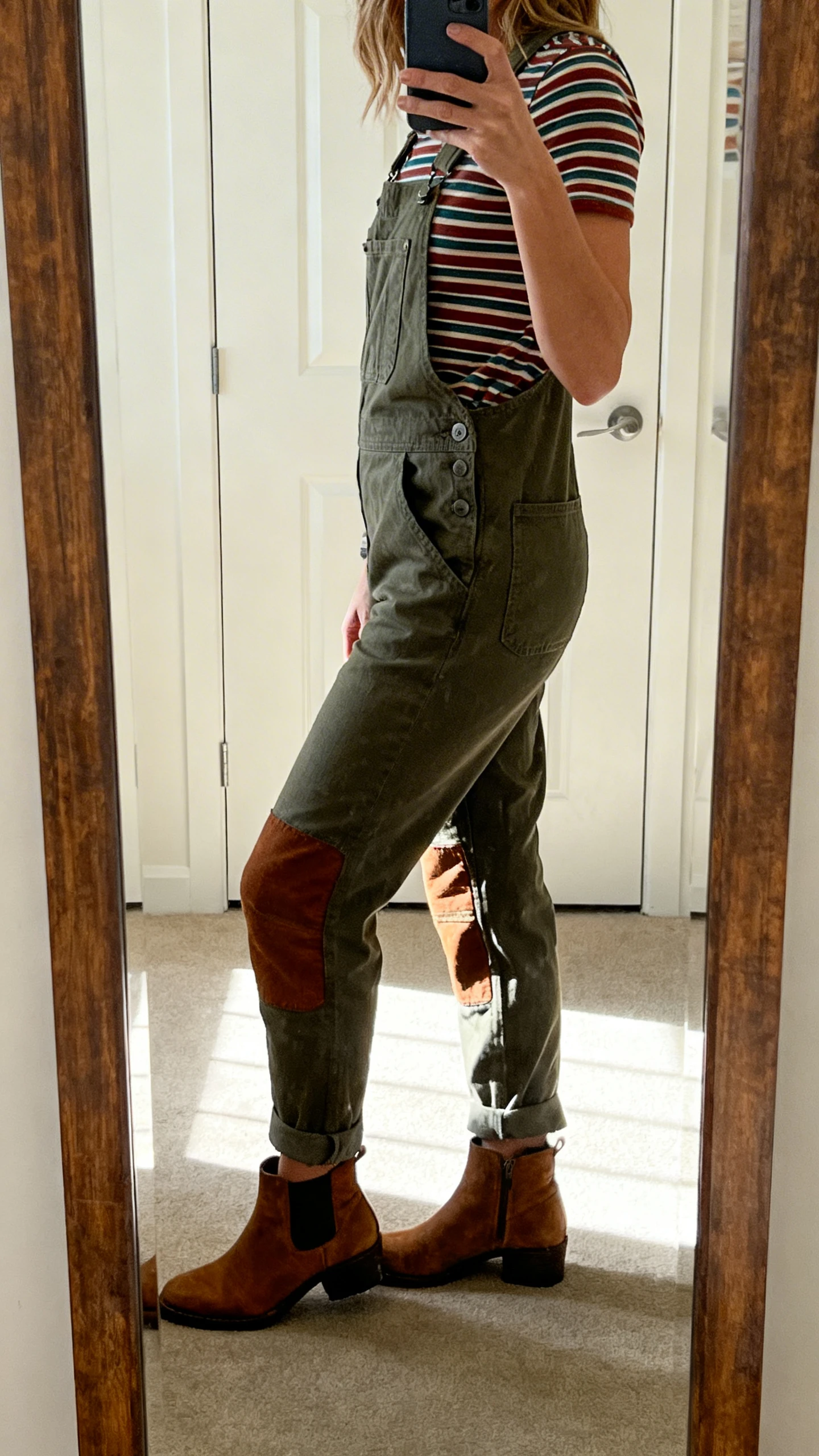 iPhone side-angle mirror selfie of a woman wearing adventure-ready utility overalls with knee patches over a striped tee, ankle boots, face not visible, full-length mirror, morning light, iPhone photo quality.