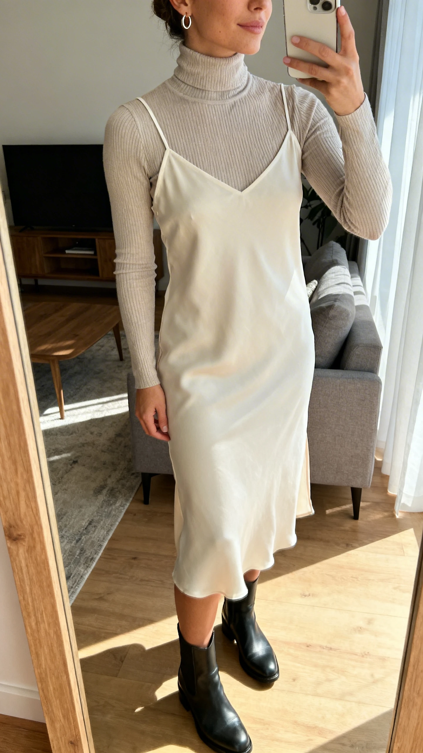 iPhone side-angle mirror selfie of a woman wearing a beginner slip dress with thin straps layered over a fitted ribbed turtleneck, ankle boots, and small hoop earrings, face not visible, living room mirror, morning natural light, iPhone photo quality.